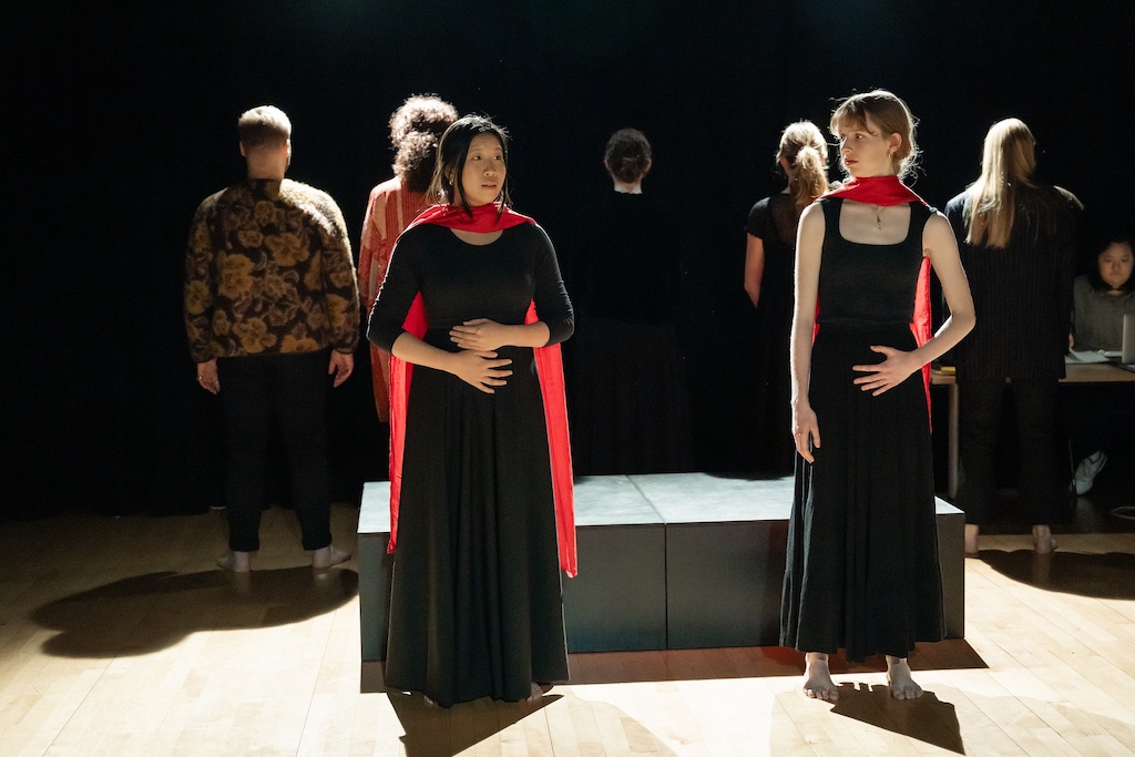 Two actors wearing black dresses and bright red capes stand on stage, slightly apart, in mid-performance.